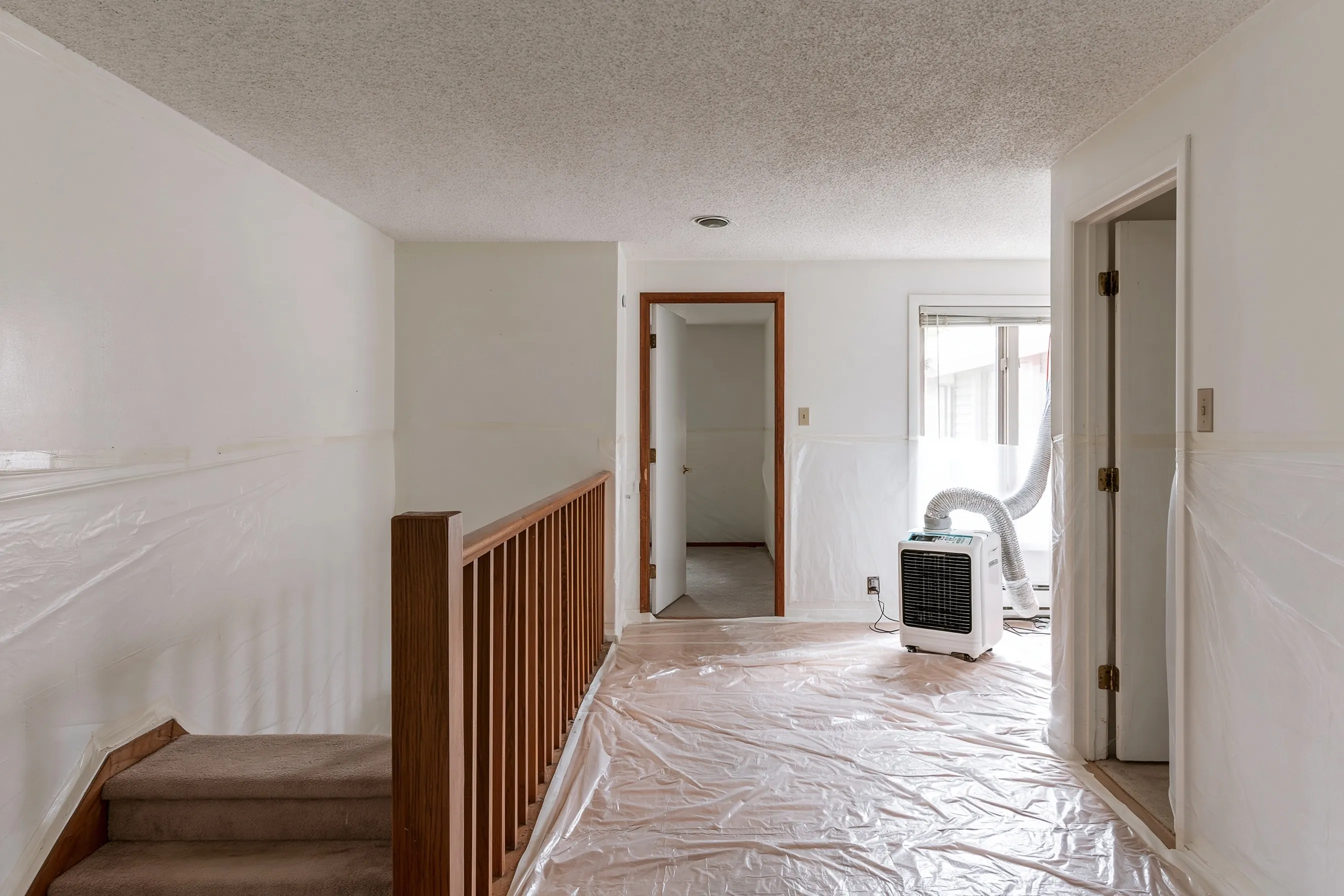 Upper floor of a house with a popcorn ceiling, plastic over the floor and walls, an air filtration unit and a half-flight of stairs in view
